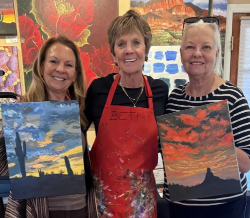 Three women holding vibrant desert landscape paintings by Beth Zink, featuring dramatic skies and silhouetted cacti.