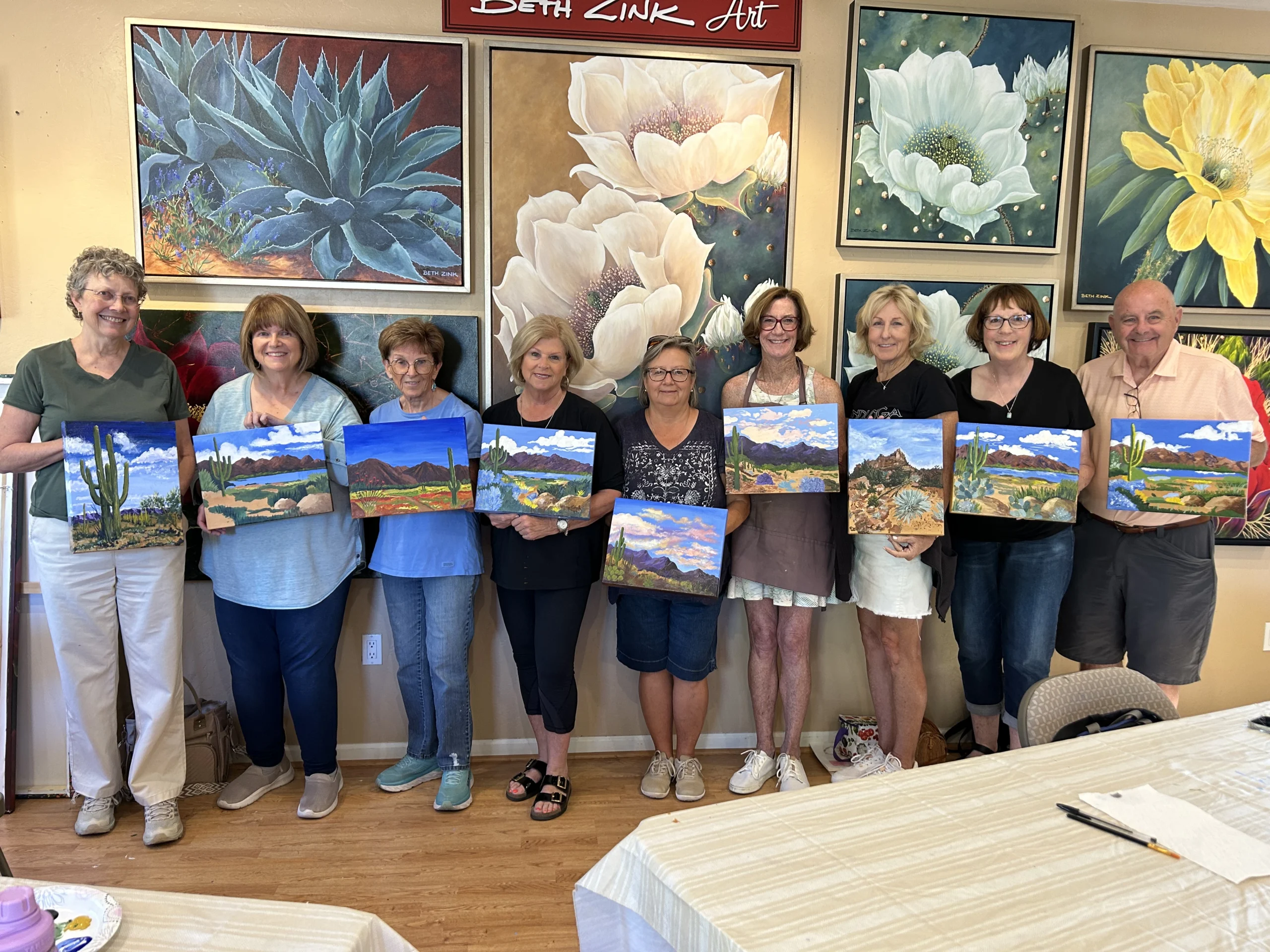 IMG_0367 Group posing with Beth Zink desert landscape paintings featuring vibrant blue skies, mountains, and desert plants.