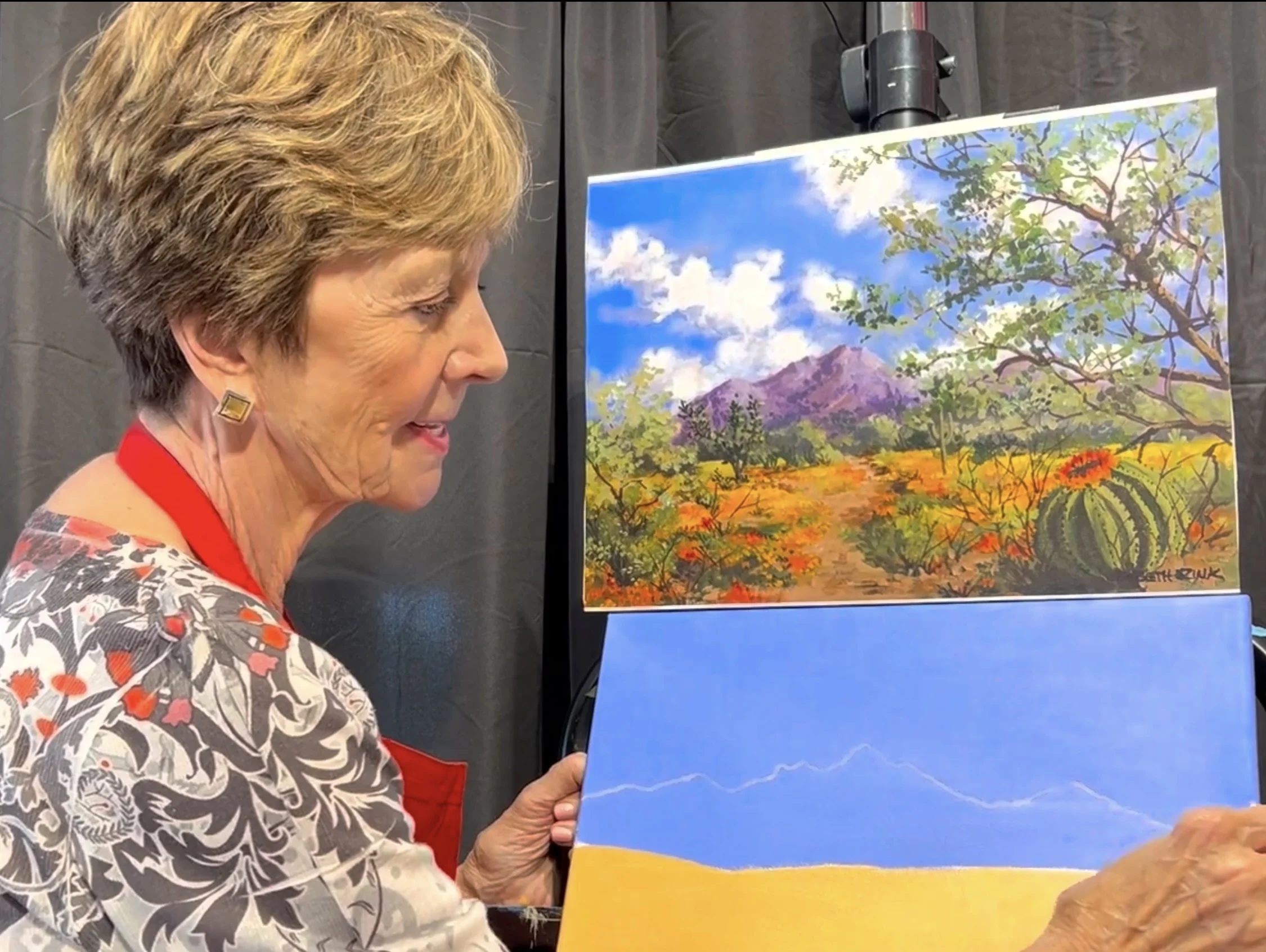 Screenshot Woman beside Beth Zink painting: desert scene with orange flowers, cacti, green shrubs, purple mountains and blue sky.