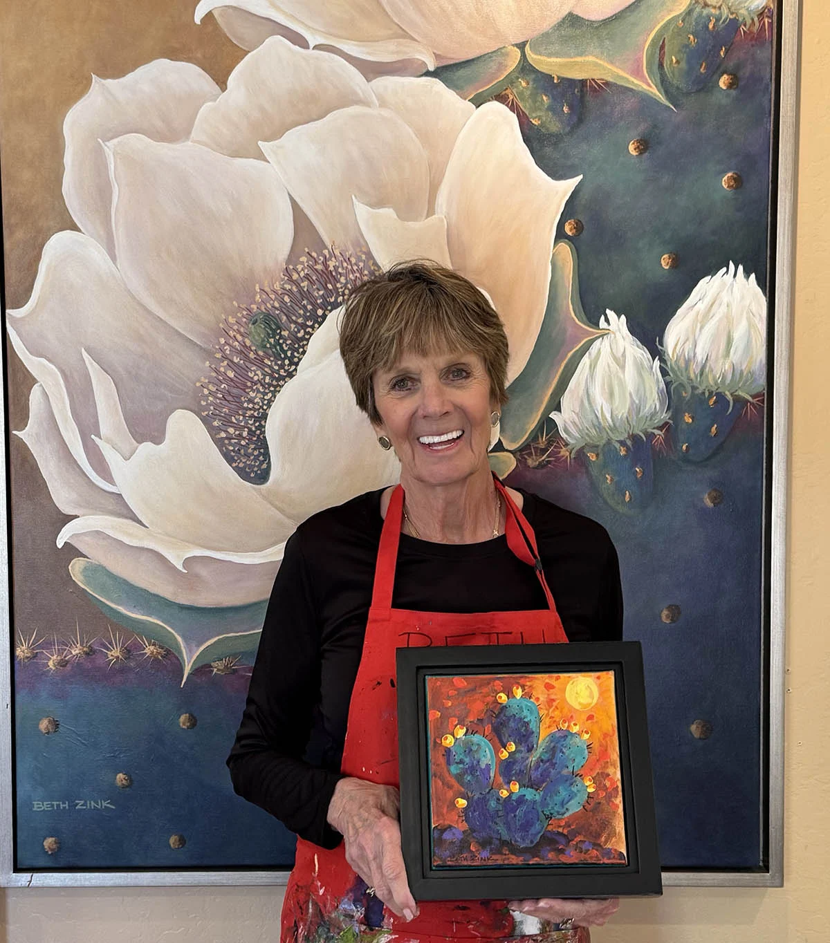 Artist Beth Zink smiles in front of her large cactus flower painting, holding a small framed artwork.