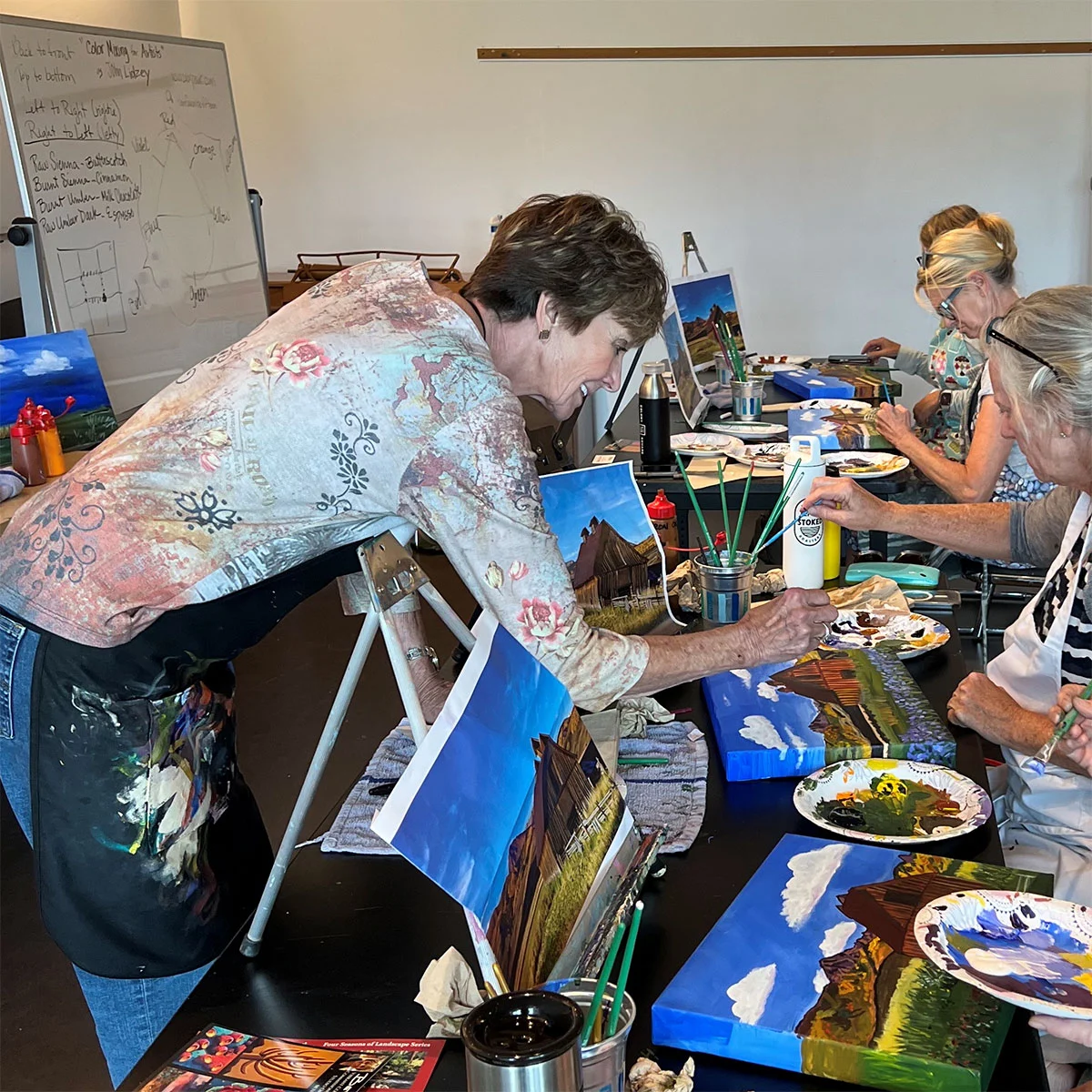 Women paint landscapes on canvases at an art class, with vibrant colors with Beth Zink guiding them.